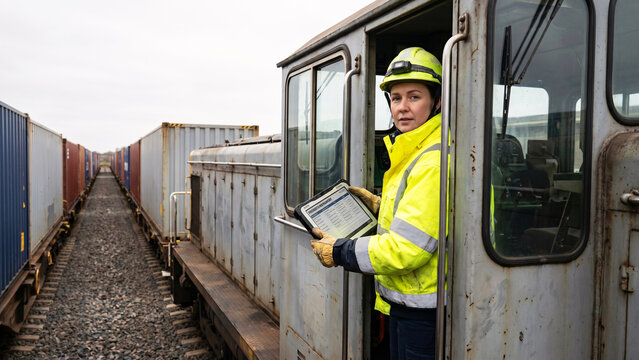 Female Freight Train Conductor in Hi-Vis Jacket Holding Industrial Tablet at Remote Rail Siding with Container Wagons, Rail Freight, Supply Chain, Logistics, Cargo