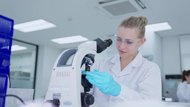 A scientist in a laboratory is focused on a microscope, looking closely at samples. The room is bright with modern equipment around her, and she wears a lab coat and gloves.