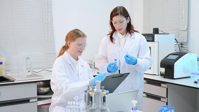 Two dedicated female scientists in lab coats working together, holding a clipboard and preparing liquid samples in test tubes during medical research