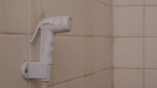 White plastic bidet hand shower sprayer mounted on clean beige square ceramic tile bathroom wall with white hose
