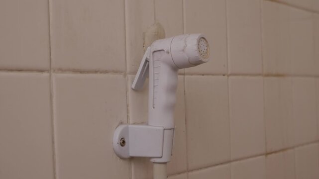 White plastic bidet hand shower sprayer mounted on clean beige square ceramic tile bathroom wall with white hose