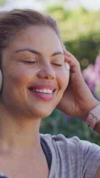 Vertical video: Smiling woman mouthing lyrics to music, adjusting white headset in park, grey top