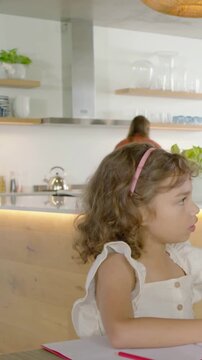 Vertical video: Drawing kid raising arm for attention as mom moving in kitchen, pencil, copy space
