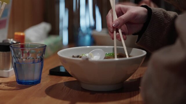 Tourist hand showing difficulty using chopsticks over ramen bowl with blue glass on table