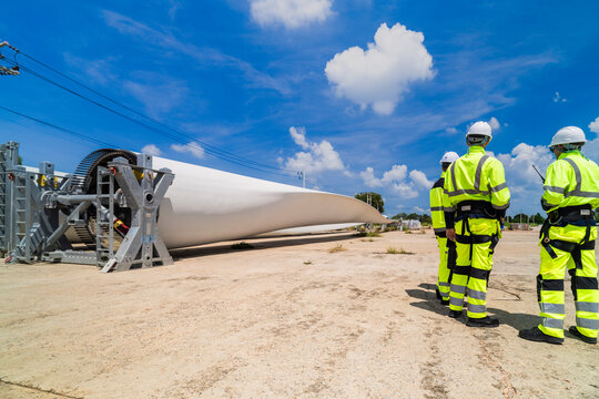 Engineers in high visibility gear assess a large wind turbine blade at a construction site under a bright blue sky, representing the implementation of renewable energy technology for sustainable.