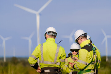 Fototapeta premium A team of engineers in high visibility safety gear stand together at a wind farm, conducting inspections and discussing renewable energy projects. Wind turbines surround them under a clear sky.