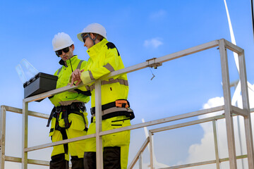 Fototapeta premium Professional wind turbine technicians in high-visibility safety gear using a laptop to monitor renewable energy operations on an elevated platform.