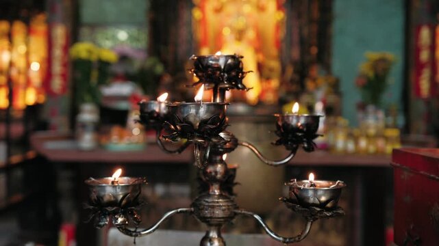 Close up of burning candles in a lotus flower shaped candelabra. A buddhist temple altar with offerings is visible in the background