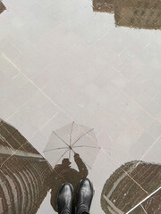 Obraz premium Reflection of person with umbrella in rain puddle outdoors. Wet city street, rainy weather, urban lifestyle, moody atmosphere.