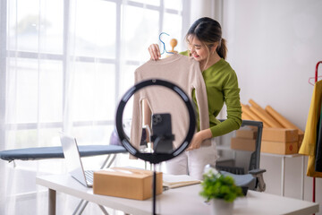 Fototapeta premium Young asian businesswoman live streaming selling clothes online via social media, showing garment to followers