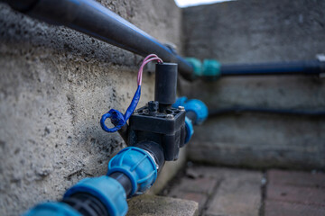 Naklejka premium Solenoid irrigation valve with wires connected to plastic pipes near concrete wall, part of outdoor garden watering system.