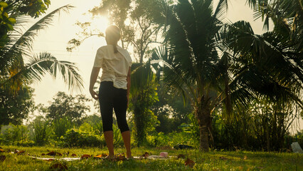 Sunlight filters through palm trees. Person stands barefoot on grassy ground. They wear loose shirt and dark pants. Silhouetted by golden light, calm and still. Perfect for tranquil © WARAPHON