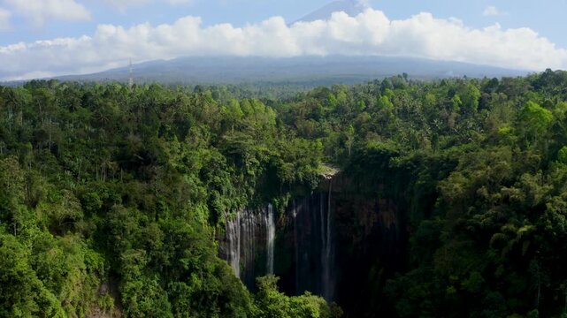 Aerial view of Grojogan Sewu Waterfall cascading down a cliff surrounded by dense tropical forest with mountains in the distance Kecamatan Pujon, Jawa Timur, Indonesia.