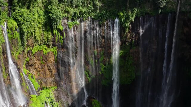 Aerial view of Grojogan Sewu Waterfall with multiple streams cascading down lush green cliffs in Kecamatan Pujon, Jawa Timur, Indonesia.