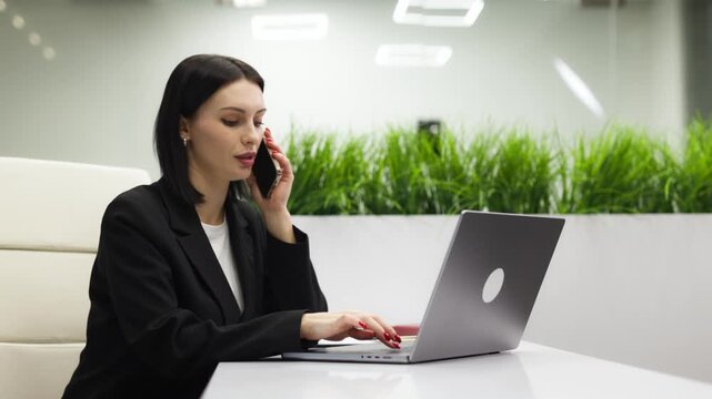 Female professional handling phone call while entering data on laptop in office. Concept customer service and business workflow.
