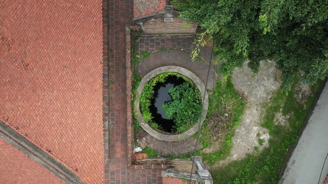 Top down drone view of ancient circular well beside traditional tiled rooftops in Duong Lam Ancient Village near Hanoi.
