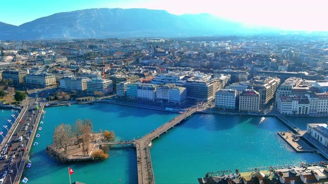 4K aerial view of Geneva with Pont du Mont-Blanc bridge, &Icirc;le Rousseau, Rh&ocirc;ne River, Saint-Pierre Cathedral spire and Sal&egrave;ve mountain horizon under winter sunlight in Switzerland.