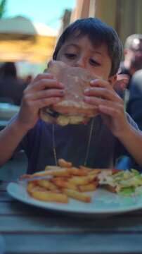 Boy eating hamburger with fries at outdoor restaurant, casual dining moment expressing appetite, enjoyment, family outing and relaxed social lifestyle