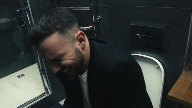 Businessman sitting on toilet in restroom showing signs of discomfort while wiping forehead with tissue. Concept digestive problems, stress and healthcare.
