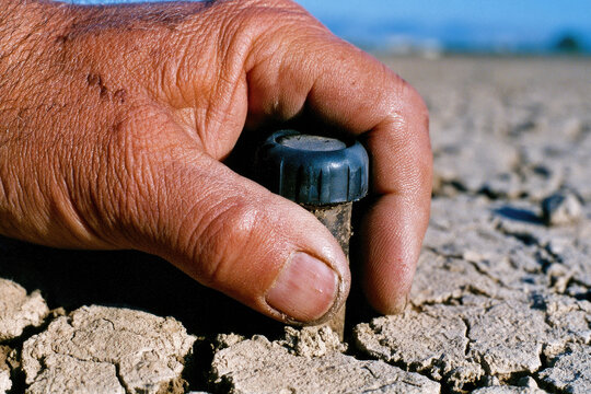 Soil moisture sensor probe in dry cracked soil with hand holding black cap, close up of smart farming tool in harsh sunlight, gritty realistic texture