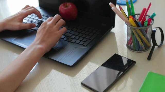Student hands typing on laptop with red apple, smartphone and colorful stationery on desk, back to school and e-learning concept, studying