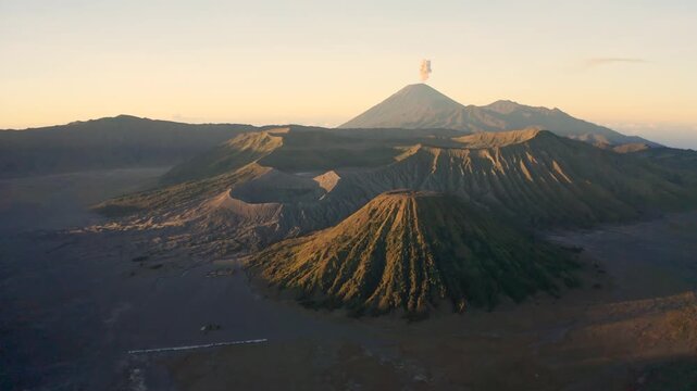 Aerial view of Mount Bromo and Mount Batok volcanic craters with Mount Semeru emitting smoke in the background during a golden sunrise over the caldera in Tosari, East Java, Indonesia.