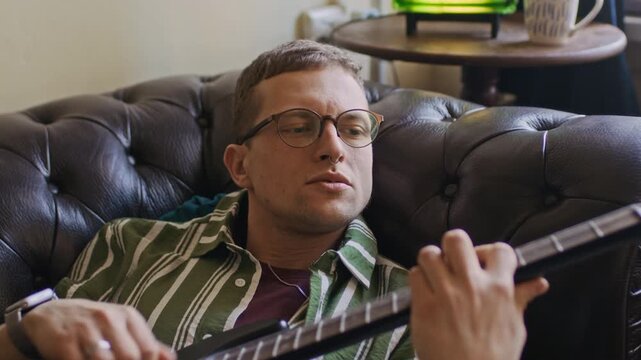 Handheld medium close up shot of tattooed Caucasian man practicing electric guitar chords while resting on sofa in apartment