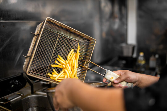 Chef dumping fresh golden french fries from a metal deep fryer basket in a commercial restaurant kitchen.