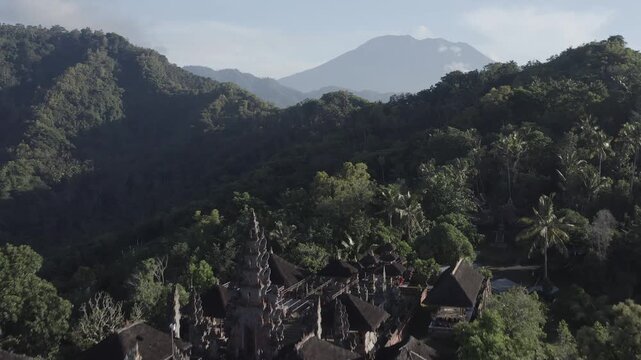 Aerial view of a traditional Balinese temple nestled in lush tropical hills with the majestic Mount Agung volcano in the background Bebandem, Bali, Indonesia.