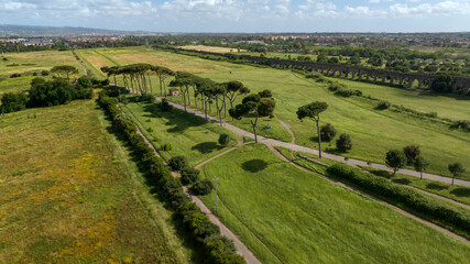 Obraz premium Aerial panoramic view of the Parco degli Acquedotti in Rome, Italy, with its iconic stone pine trees and green meadows stretching toward the distant city skyline under a clear blue summer sky.
