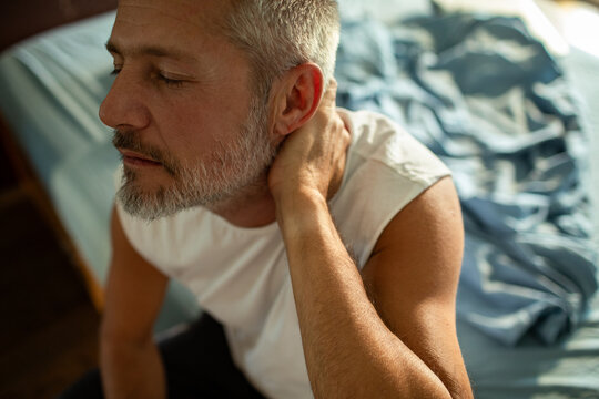Middle-aged man with neck pain sitting on bed at home