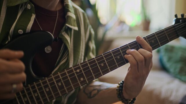 Handheld close up shot of tattooed Caucasian man with nose piercing playing guitar while practicing chords and singing at home