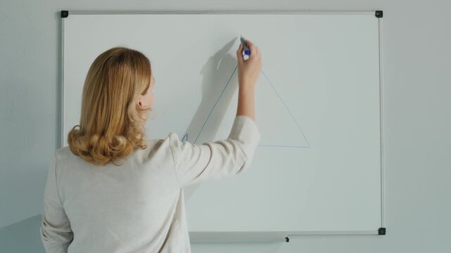 Teacher drawing equilateral triangle on whiteboard during mathematics lesson in classroom with blue marker.
