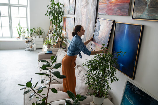 Talented woman painter hanging new finished artwork canvas on wall at home loft gallery, showcasing skills and love for art. Successful female artist arranging own exhibition in studio living room.