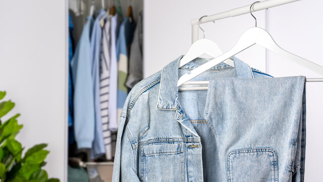 Light blue denim jacket and jeans on white wooden hangers on clothing rack, open closet on background. Concept of wardrobe organization, minimalist clothing and casual fashion lifestyle. Copy space