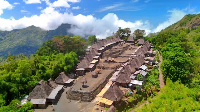 Aerial view of Bena Traditional Village with iconic thatched roofs surrounded by dense tropical greenery and misty mountains under a blue sky in Kecamatan Jerebuu, Nusa Tenggara Timur, Indonesia.