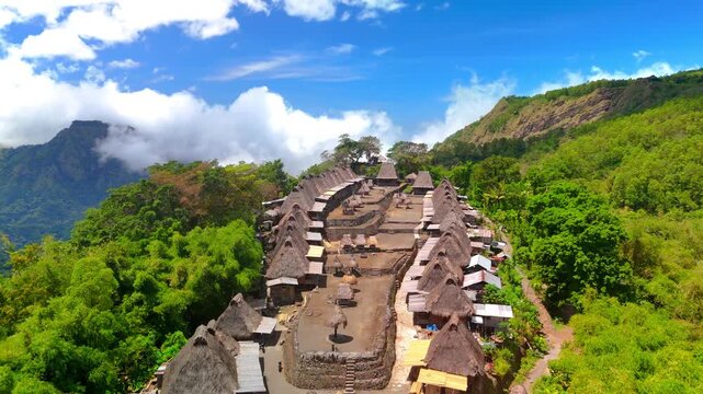 Aerial view of Bena Traditional Village with its iconic thatched roof houses nestled in lush green hills under a blue sky in Kecamatan Jerebuu, Nusa Tenggara Timur, Indonesia.