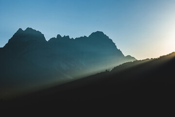 Obraz premium sole che tramonta dietro alle montagne, sagome scure delle catene montuose che vengono messe in risalto dalla luce del sole in un cielo sereno