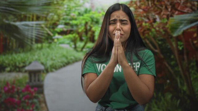 Woman volunteer in green t shirt with hands pressed in prayer gesture and pleading facial expression on a garden path in forest; hope plea.