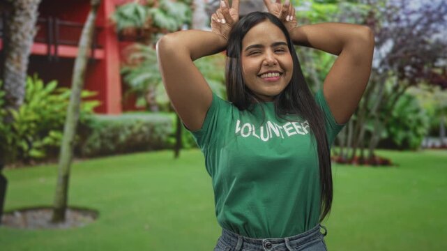 Volunteer woman in green tshirt smiling with long hair and arms raised, making bunny ears with fingers in a manicured garden by a red building; community joy.