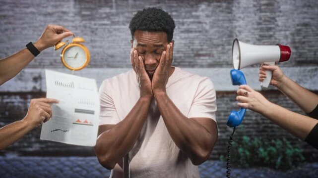 Man holding face with hands while an alarm clock, paper charts, a telephone handset and megaphone are waved around him in studio; stress overwhelm choices.