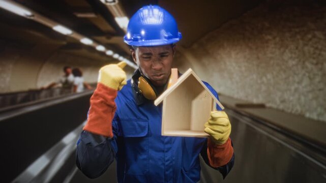 Man builder wearing blue hardhat and yellow ear protectors holding a small wooden house while raising a gloved hand in building; pride duty.