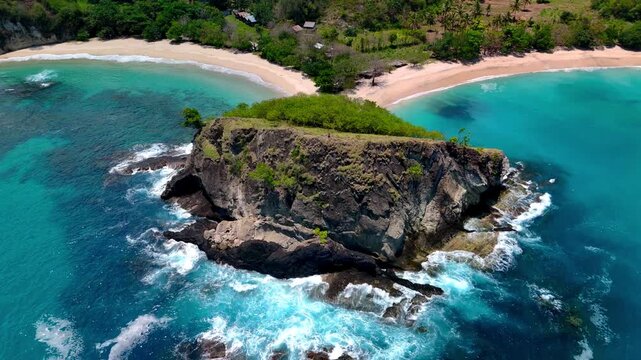 Aerial view of Koka Beach featuring a large rocky cliff separating two white sand bays with turquoise water and lush green vegetation in Paga, East Nusa Tenggara, Indonesia.