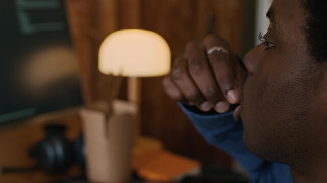 Handheld shot of male African American freelance developer programming on computer with script on screen and eating french fries while working from home