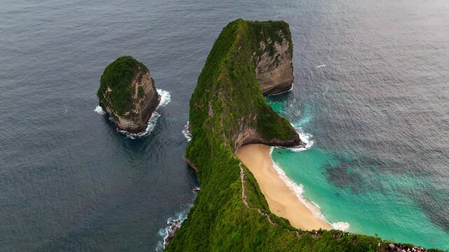 Aerial view of Kelingking Beach featuring the famous T-Rex shaped cliff, turquoise ocean waves, and tourists gathered at the viewpoint Nusa Penida, Bali, Indonesia.