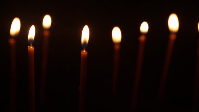 Hand holding burning candle moves along a row of seven unlit church candles, sequentially touching each wick and lighting them one by one until all burn together in warm golden light.
