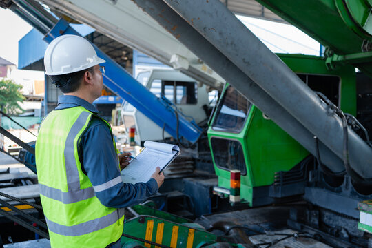 Engineer in hard hat reviewing crane inspection checklist while looking up at boom and operator cab in equipment yard