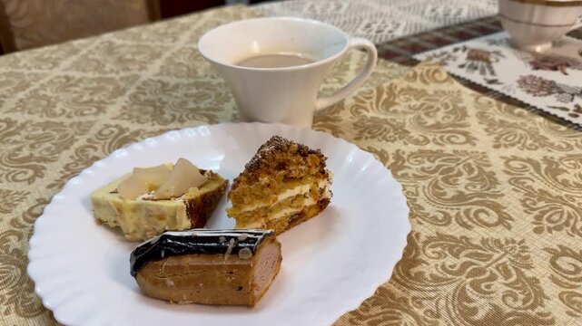 Plate with coffee and pastries on table, white plate featuring chocolate eclair and cake pieces next to porcelain mug, ornate gold