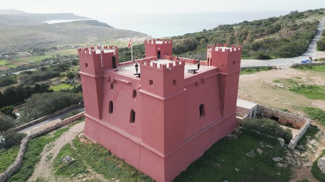 Cinematic aerial shot rising smoothly to reveal the red stone walls of Saint Agatha's Tower, also known as the Red Tower in Malta.