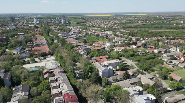 Drone rotating shot showing town panorama of Vrbas, Serbia with lush green trees and urban architecture under a blue sky.